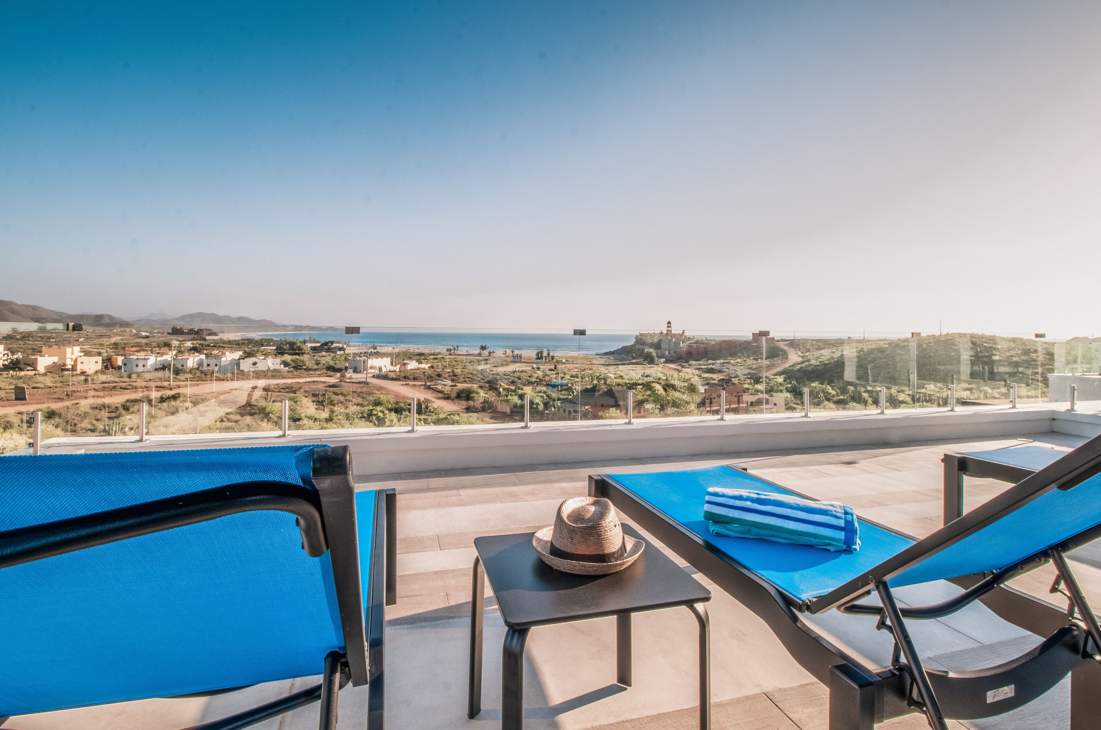 Un balcon avec deux fauteuils bleus, une petite table sur laquelle repose un chapeau, et une vue sur la mer et les bâtiments éloignés.