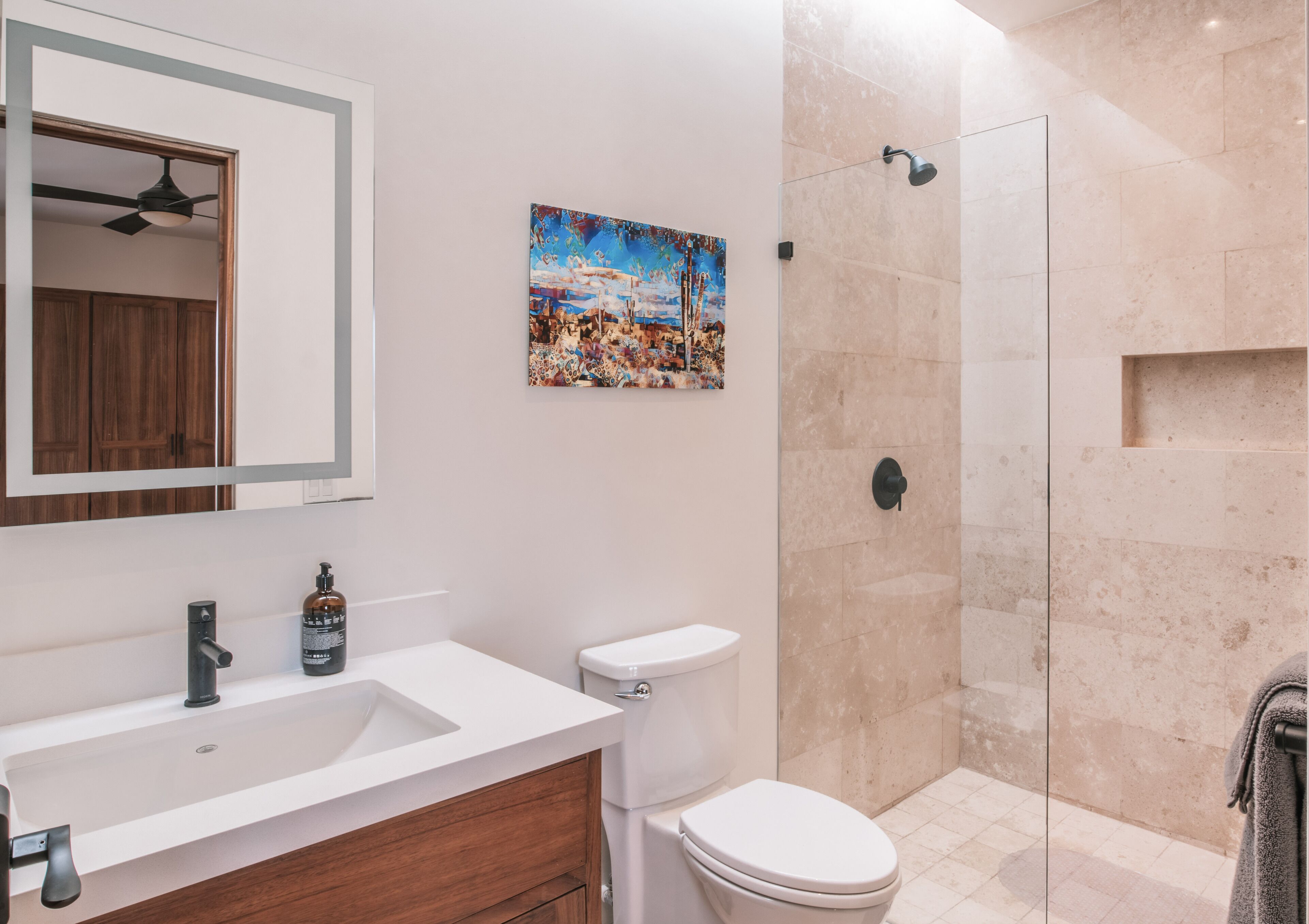 Un baño moderno con lavamanos blanco, tocador de madera, inodoro, cabina de ducha con mampara de vidrio y ventilador de techo.