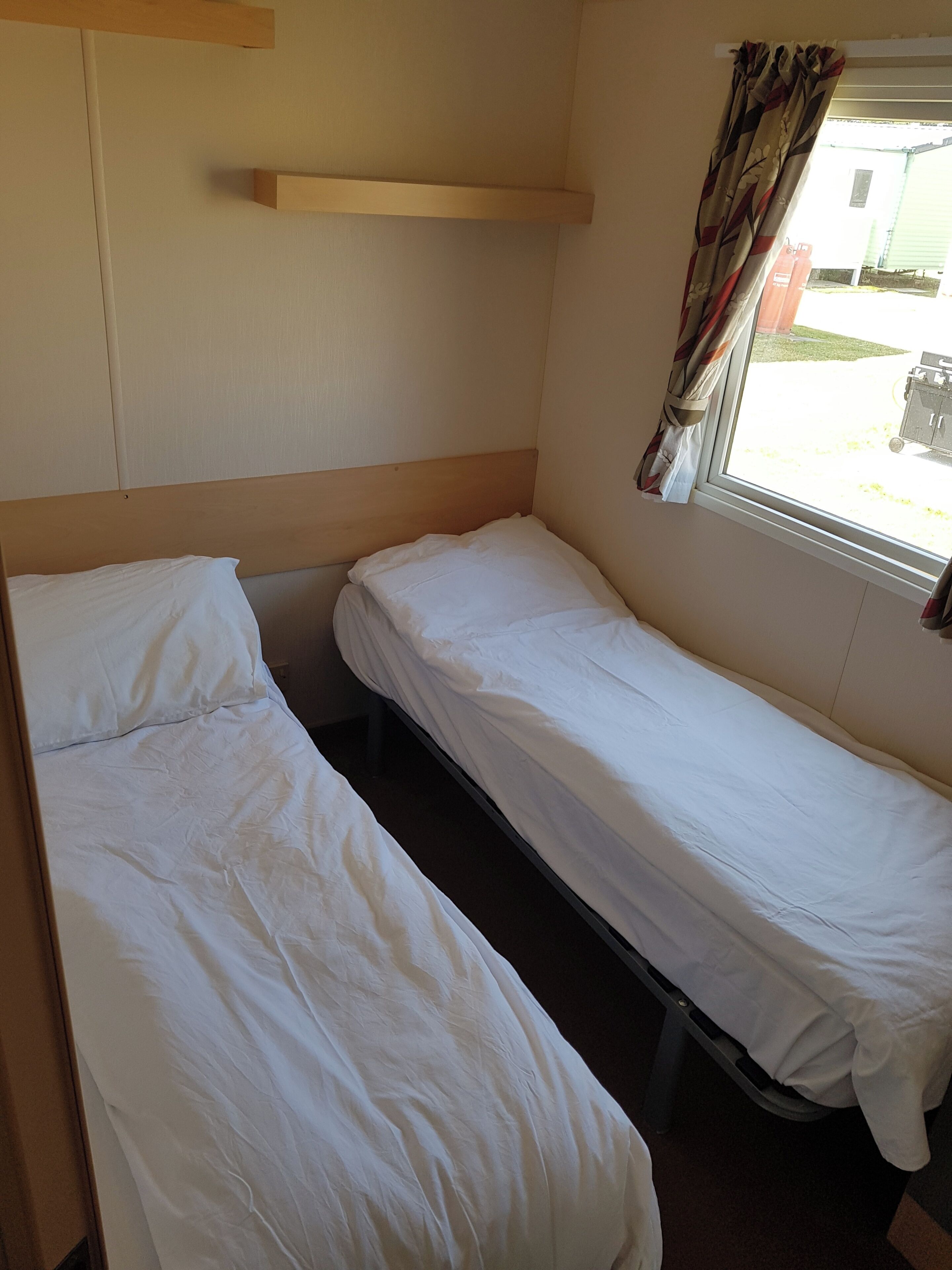 Un petit logement composé d’une seule pièce, avec deux lits simples, une fenêtre avec des rideaux et une étagère en bois au-dessus des lits.