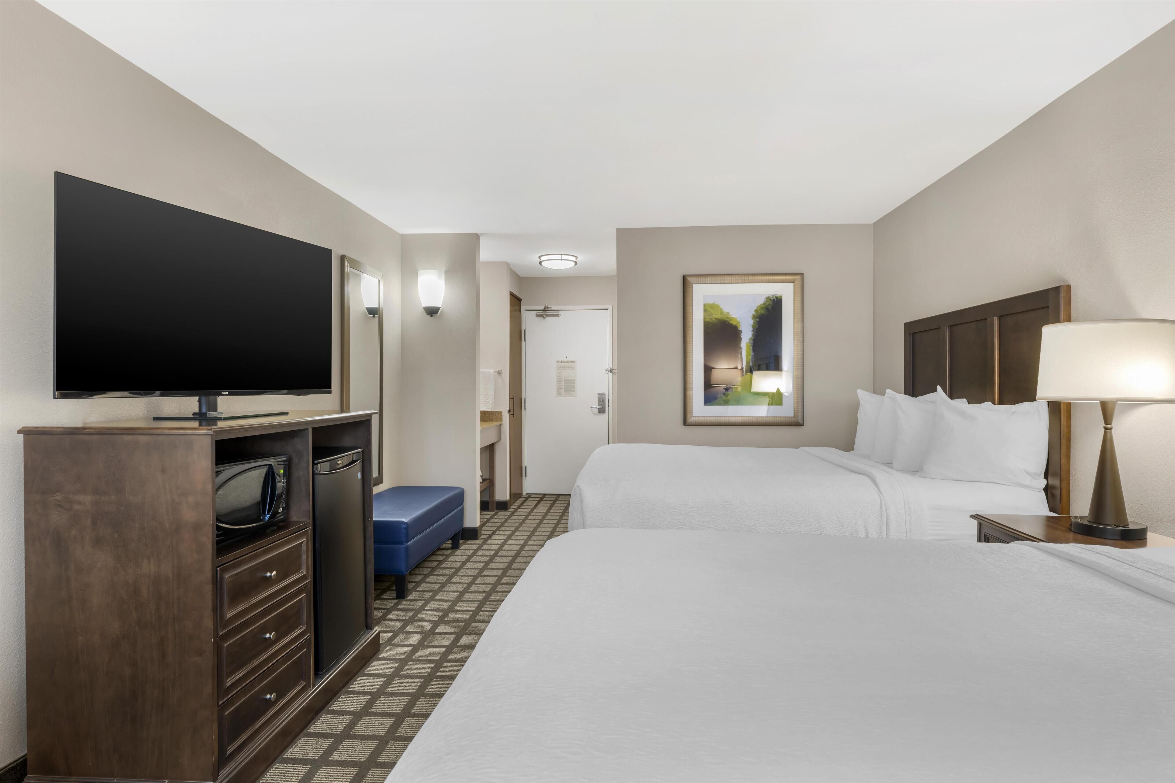 A hotel room with a flat-screen TV, a minibar, two beds, a desk, and a lamp.