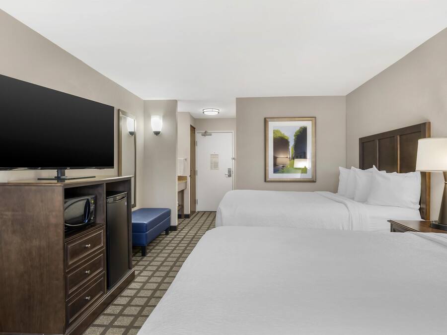 A hotel room with a flat-screen TV, a minibar, two beds, a desk, and a lamp.
