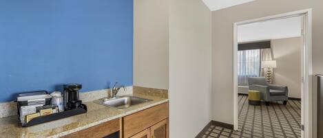 A hotel kitchen with a sink, coffee maker, and cabinets. There is a patterned carpet and a chair in the dining area.