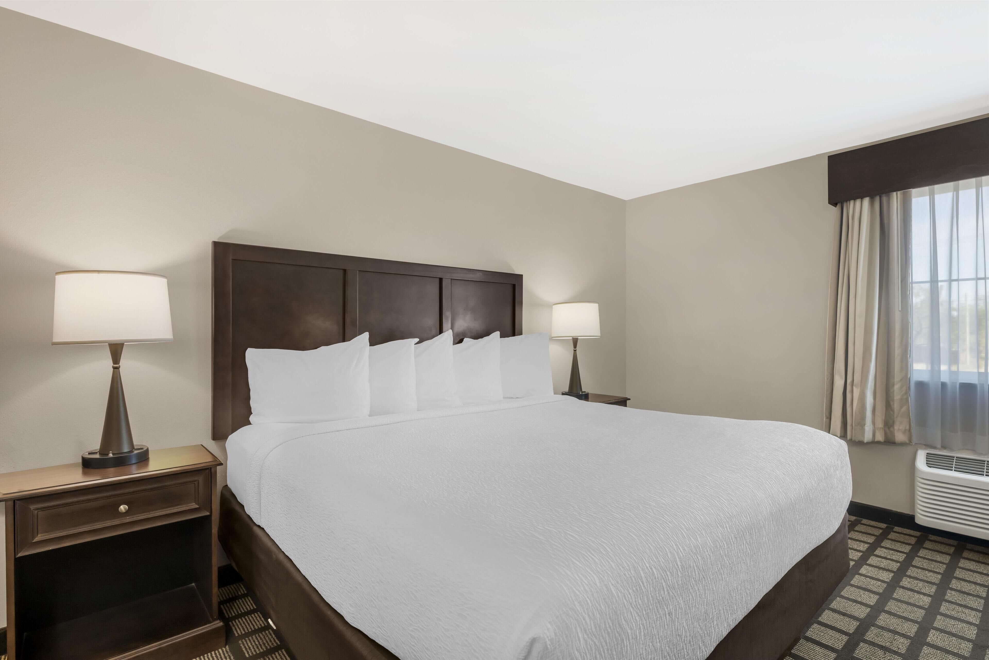 A hotel room with a large bed, two bedside tables with lamps, a window with curtains, and a wall-mounted air conditioner.