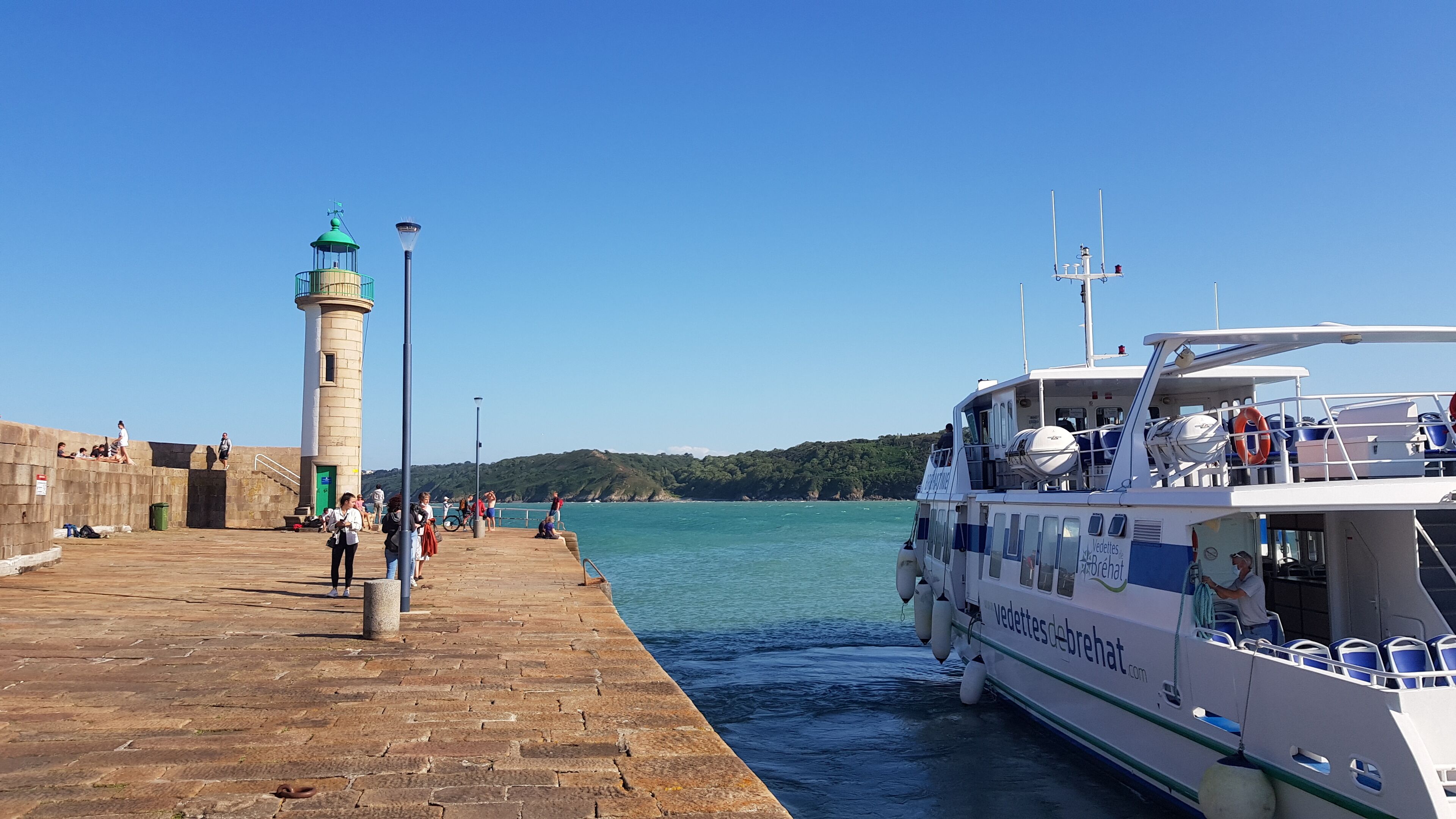 Un muelle con un faro, una embarcación amarrada al final, y gente caminando por la pasarela.