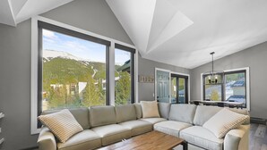 Ein modernes Wohnzimmer mit einem großen Ecksofa, einem Holztisch und großen Fenstern, die Blick auf die Berge bieten.