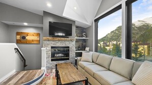Ein modernes Wohnzimmer mit Kamin aus Stein, einem großen Fenster mit Blick auf die Berge, einem Holztisch und einem beigen Sofa.