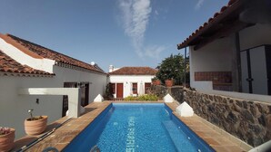 Une piscine entourée d’une terrasse carrelée, elle-même bordée de bâtiments blancs aux toits recouverts de tuiles rouges, et d’un mur de pierre d’un côté.
