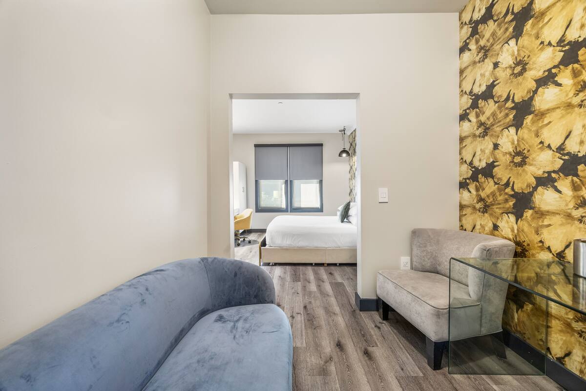 A modern hotel room with a grey sofa, a glass table, and a bed with a grey headboard.