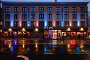 Un hotel con facciata moderna, diversi negozi al piano terra e strada bagnata che riflette luci colorate.