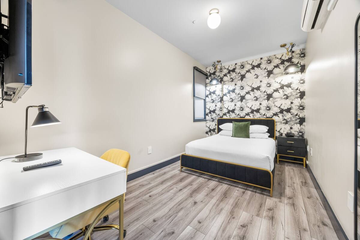 A bedroom with a bed, a desk, a chair, a nightstand, and a wall-mounted mirror.
