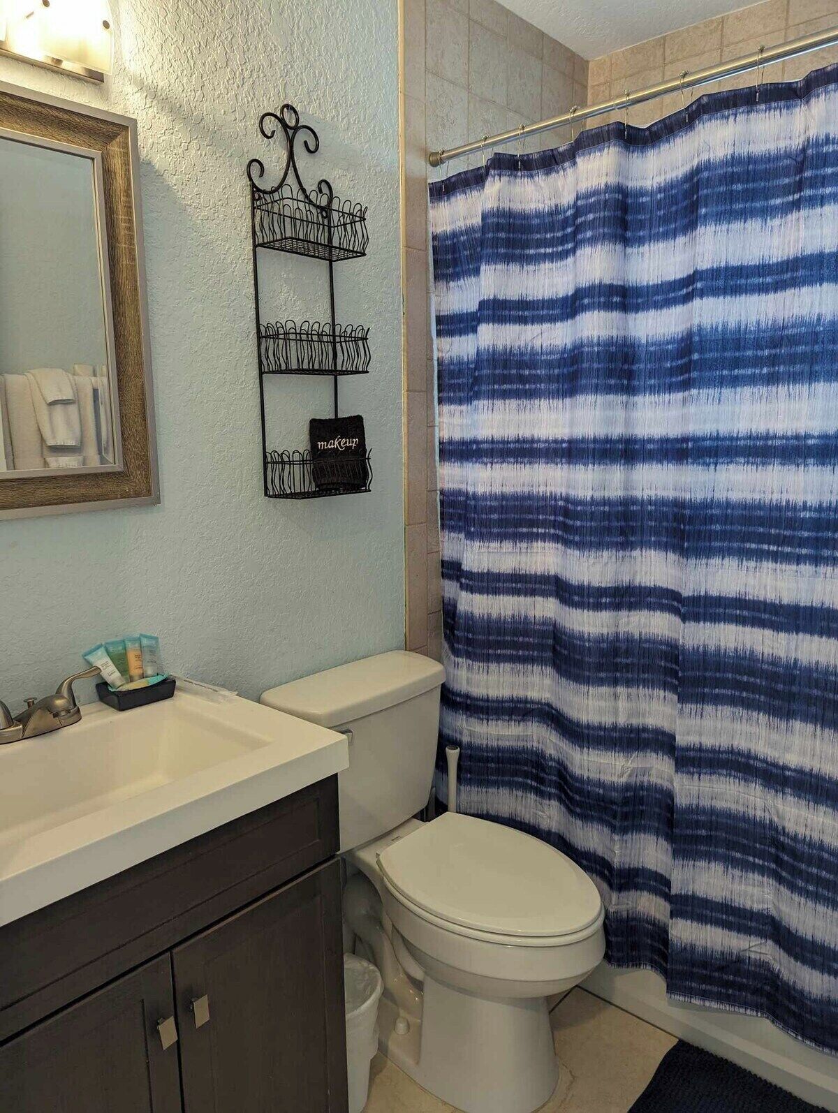 A bathroom with a white toilet, a sink with a mirror, a towel rack with a basket, and a blue-striped shower curtain.