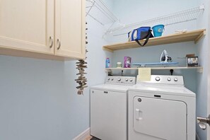 Un espace buanderie comprenant deux machines à laver blanches, une étagère avec des produits de nettoyage et un étendoir placé au-dessus.