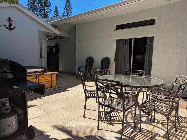 Un patio exterior con una mesa redonda de vidrio y sillas de metal, una parrilla y un armario de madera.