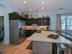 Una cocina con armarios oscuros, un refrigerador de acero inoxidable, una isla de encimera de granito y un comedor con sillas metálicas.