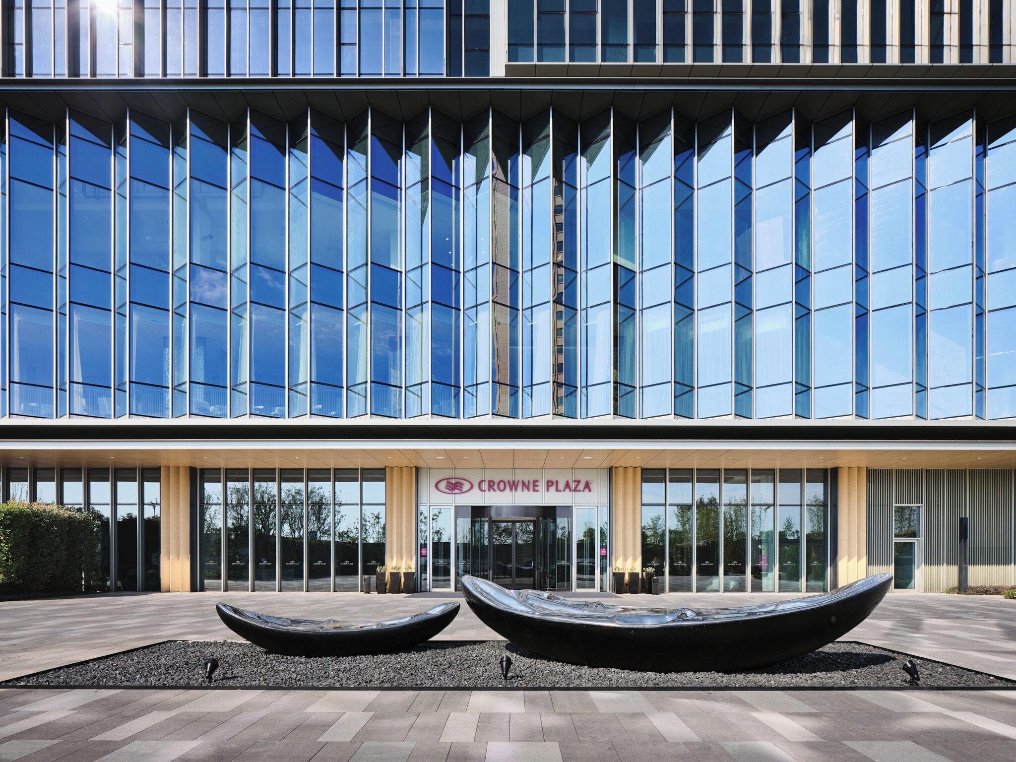 Een modern gebouw met een glazen gevel, een bord met de tekst 'CROWNE PLAZA' en twee grote zwarte plantenbakken op de voorgrond.