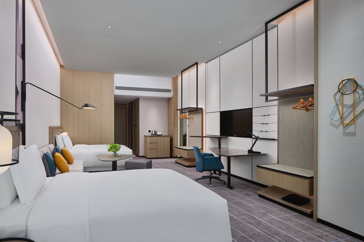 A modern hotel room with a large bed, a desk, a chair, a TV, and a wardrobe.