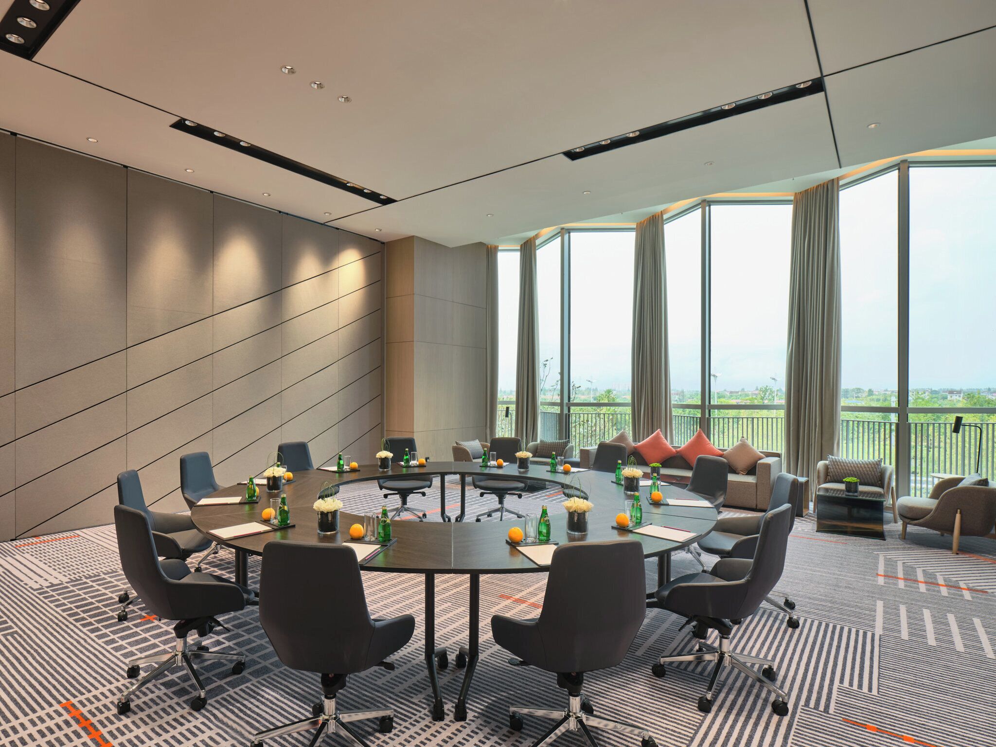 A modern conference room with a round table, multiple chairs, and a view of greenery through large windows.