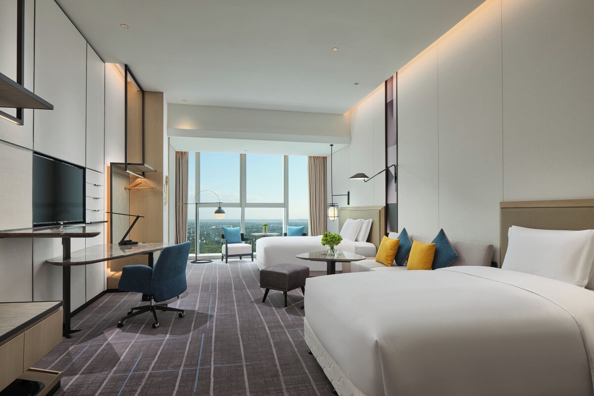 Een moderne hotelkamer met een groot bed, een bureau met een flatscreen televisie, een comfortabele fauteuil en uitzicht op de stad door grote ramen.