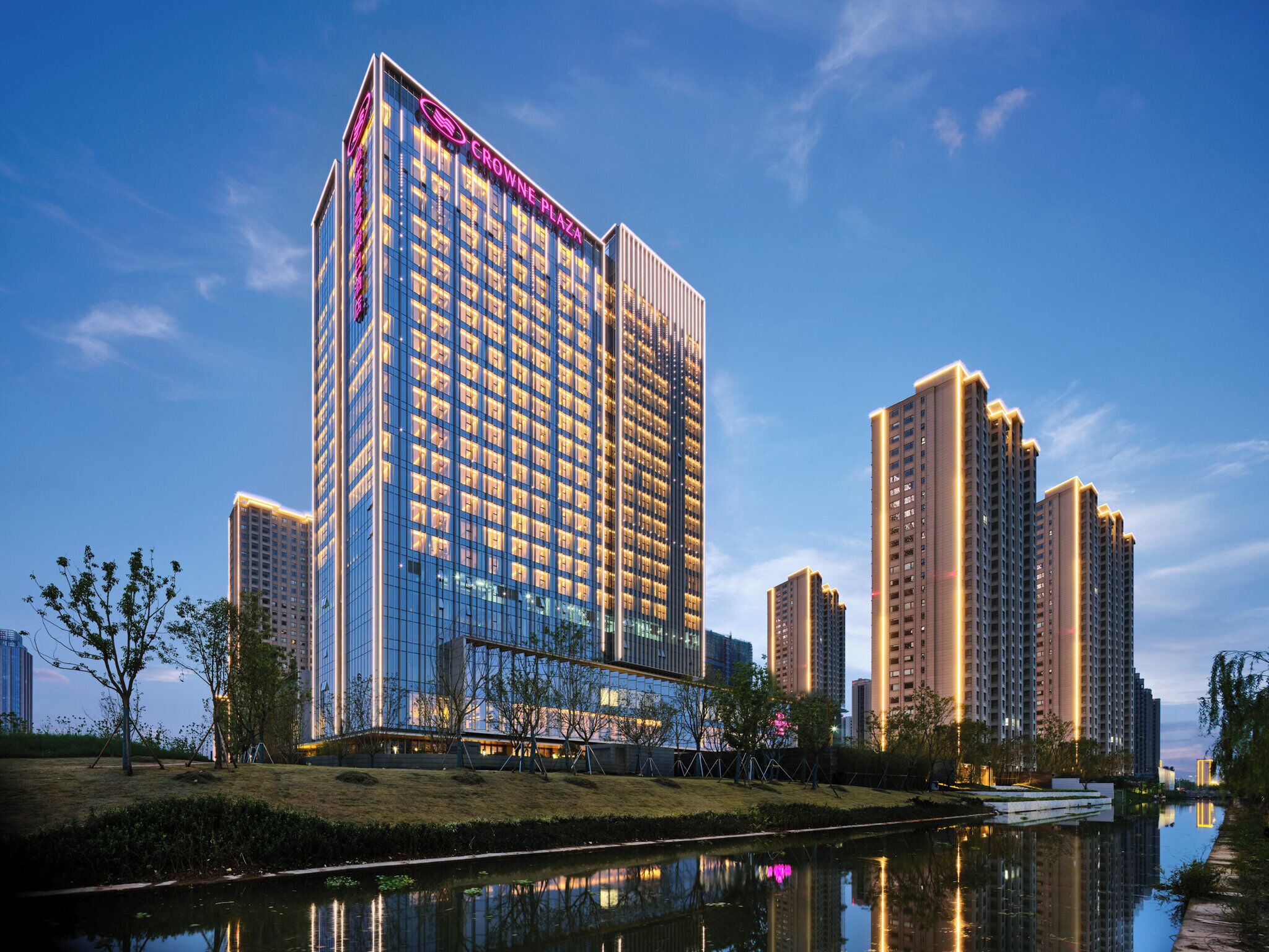 Een hoog hotel met een moderne glazen gevel, omgeven door andere hoge gebouwen, en een rustig waterlichaam dat de gebouwen weerspiegelt.