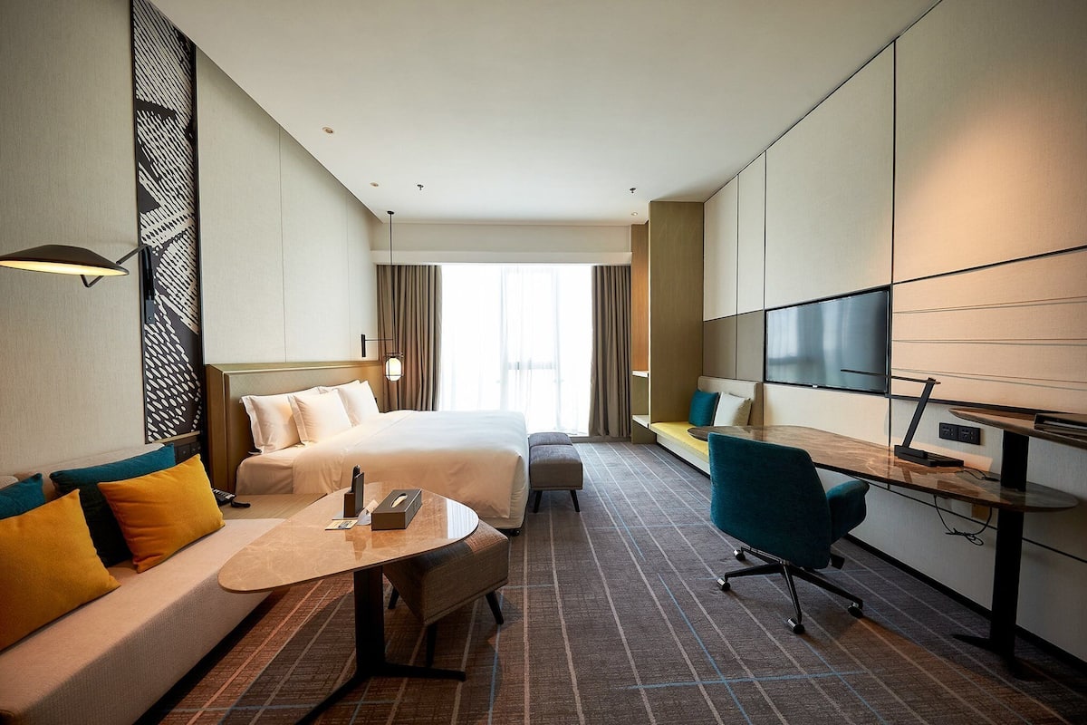 A hotel room with a bed, a sofa, a desk, a chair, a TV, and a large window.