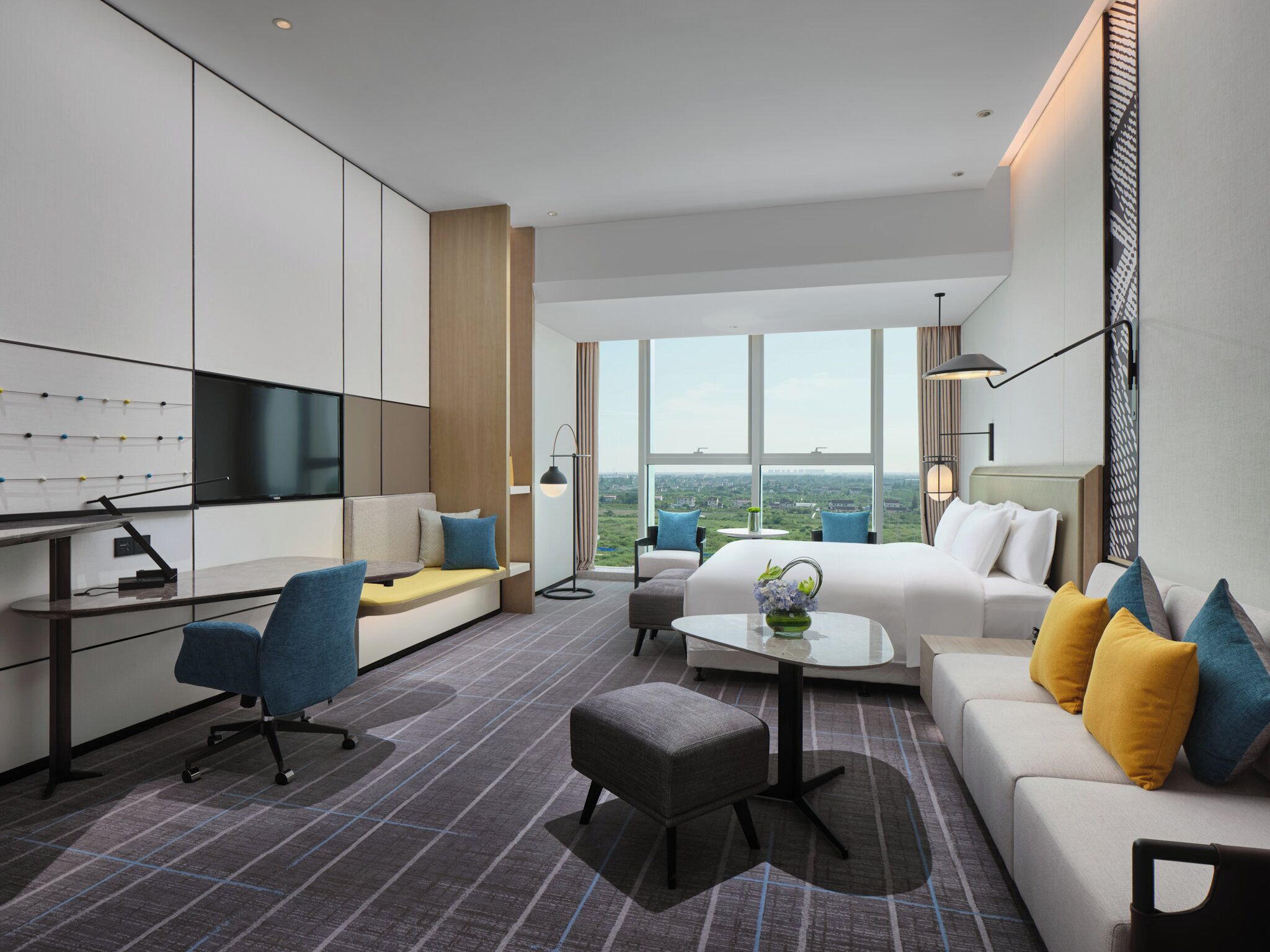 Una habitación de hotel moderna con un ventanal grande, un televisor de pantalla plana, un sofá, una mesita de centro y un escritorio con silla.
