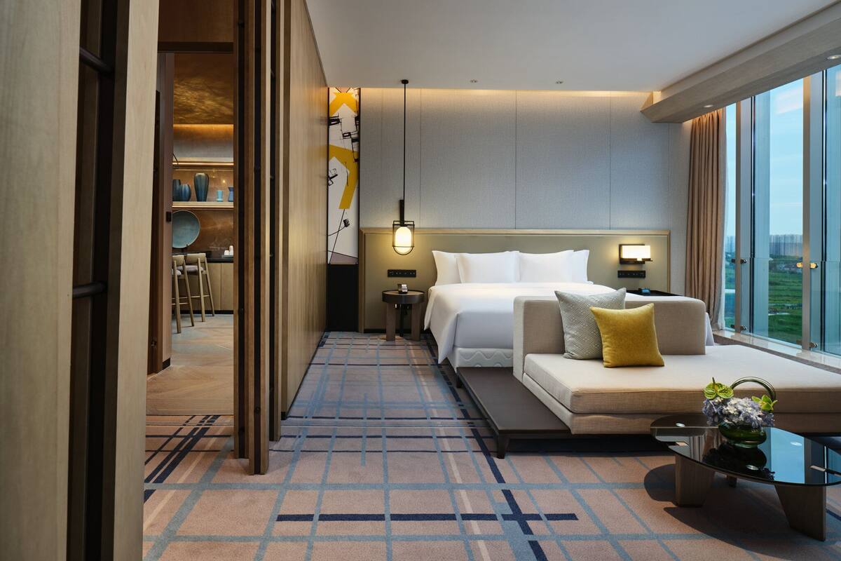 A modern hotel room with a bed, a sofa, a small table with a vase, and a view of the city.