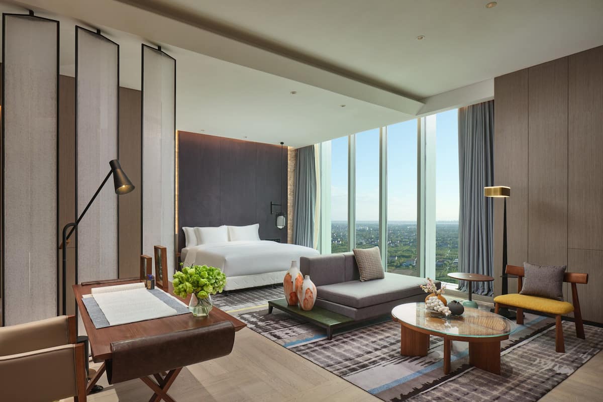 A modern hotel room with a large bed, a sofa, a dining table, and a view of the city through large windows.