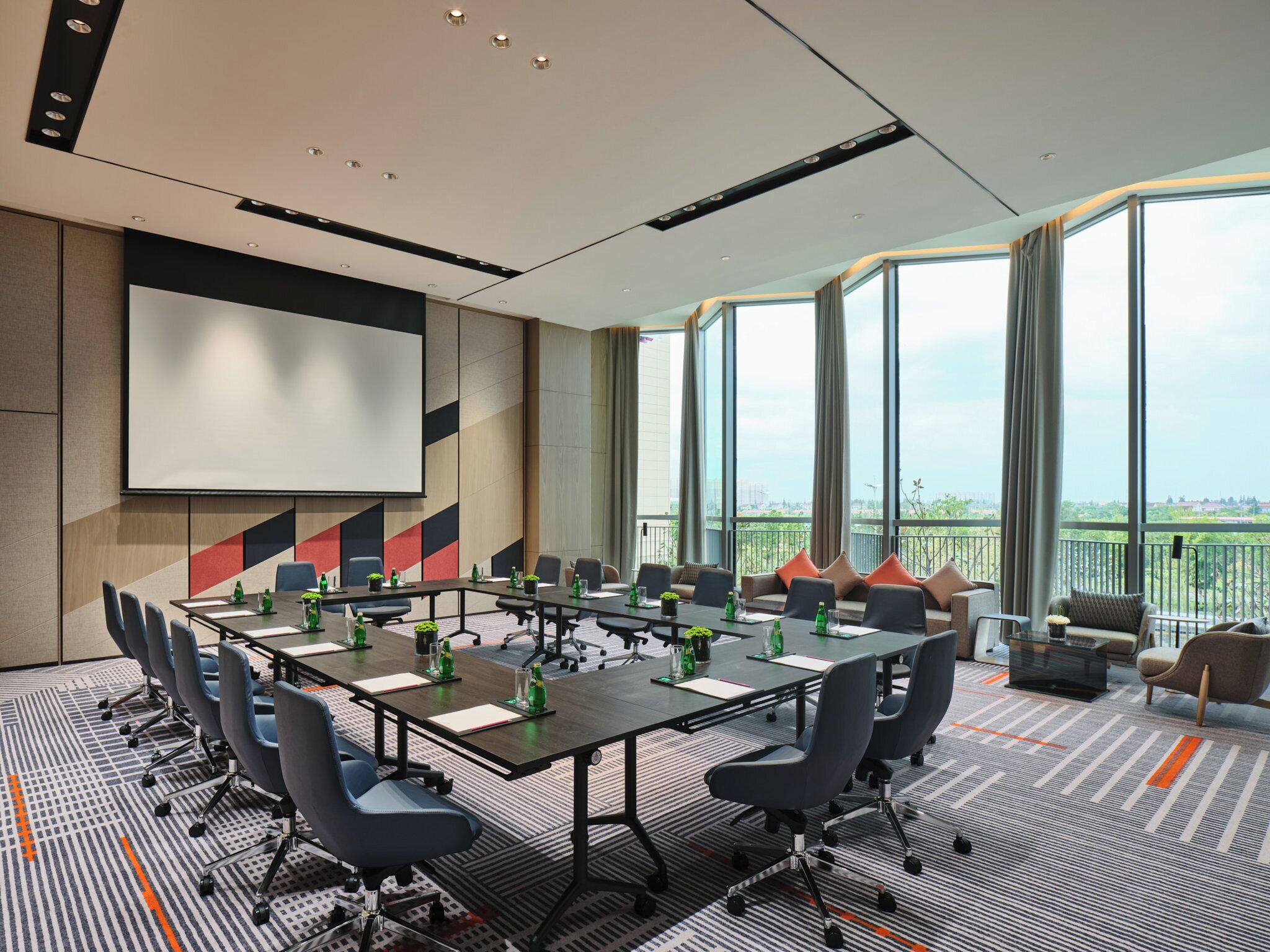 Un salón de conferencias con una mesa grande, sillas, una pantalla de proyector y vistas de la ciudad a través de amplias ventanas.
