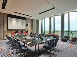 Un salón de conferencias con una mesa grande, sillas, una pantalla de proyector y vistas de la ciudad a través de amplias ventanas.