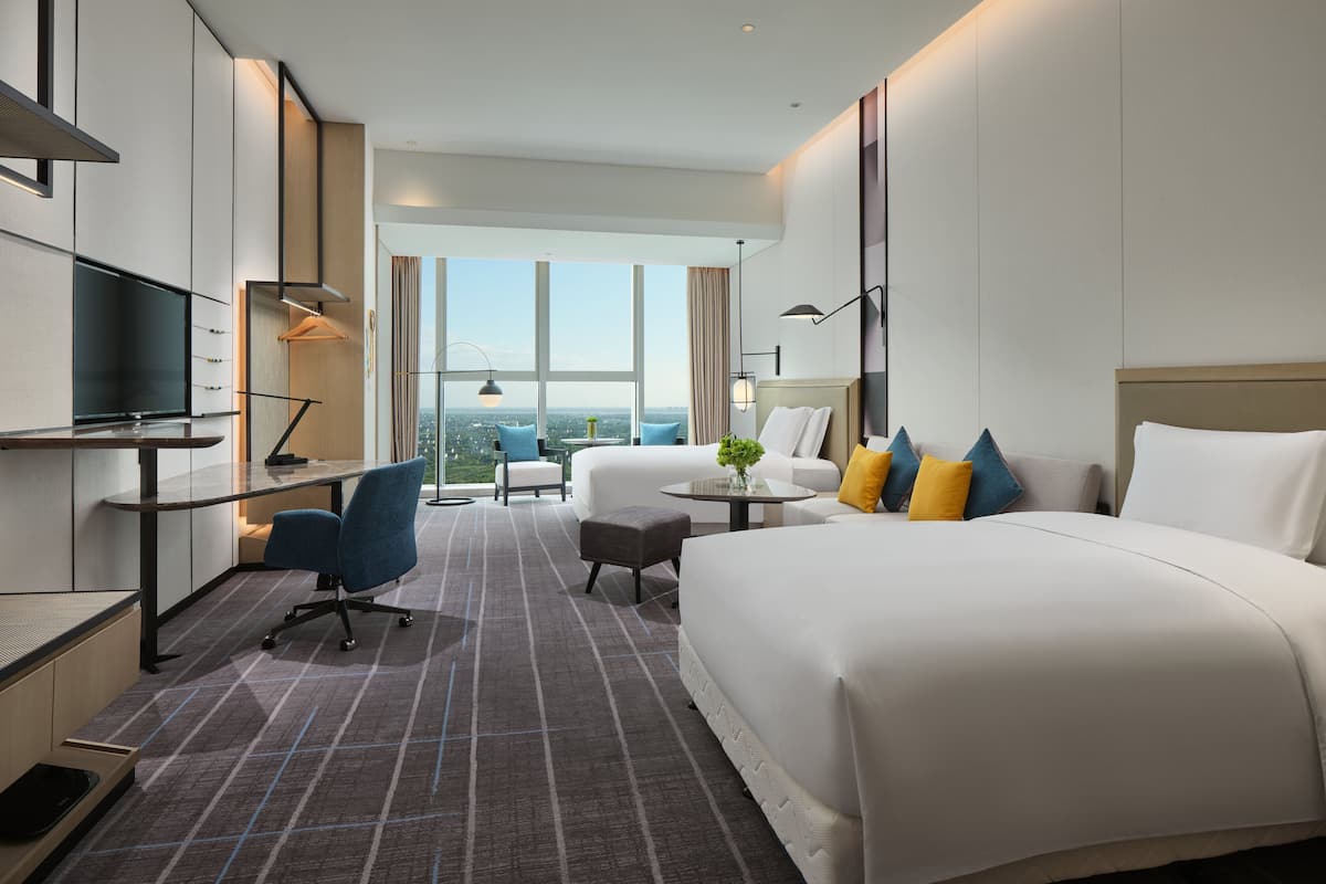 A modern hotel room with a large bed, a desk with a flat-screen TV, a comfortable armchair, and a view of the city through large windows.