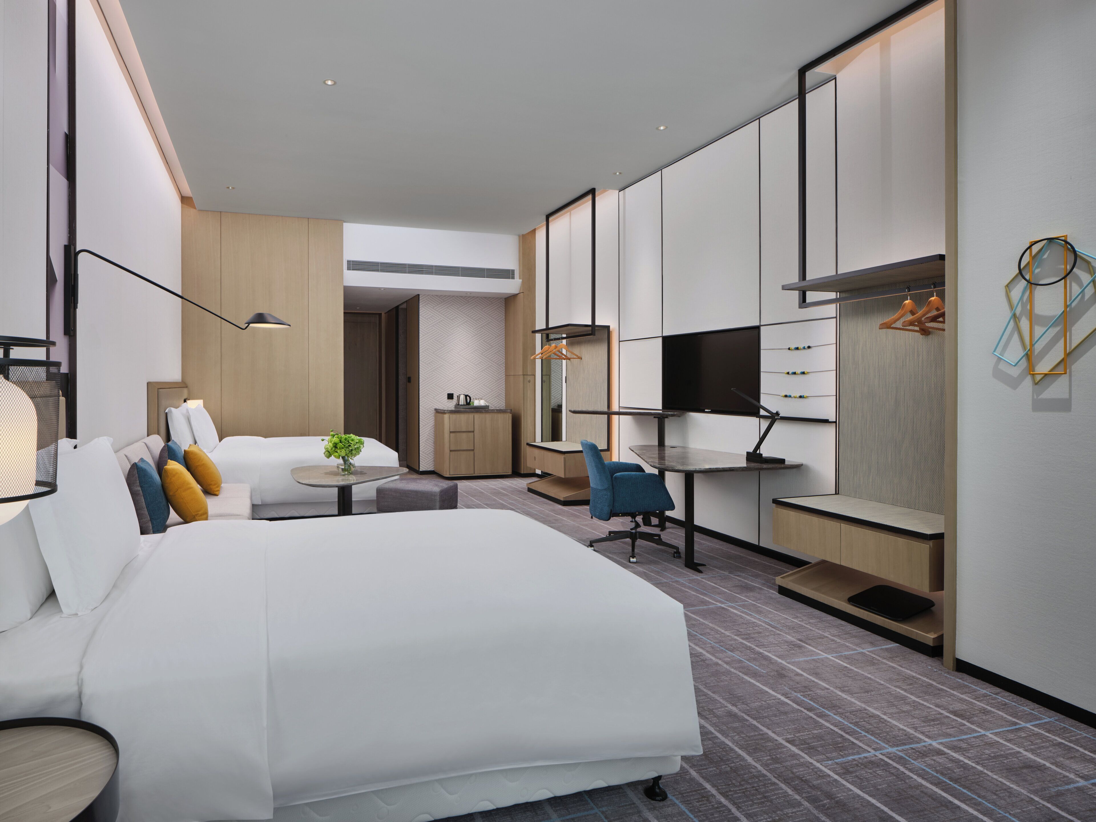 A modern hotel room with a large bed, a desk, a chair, a TV, and a wardrobe.