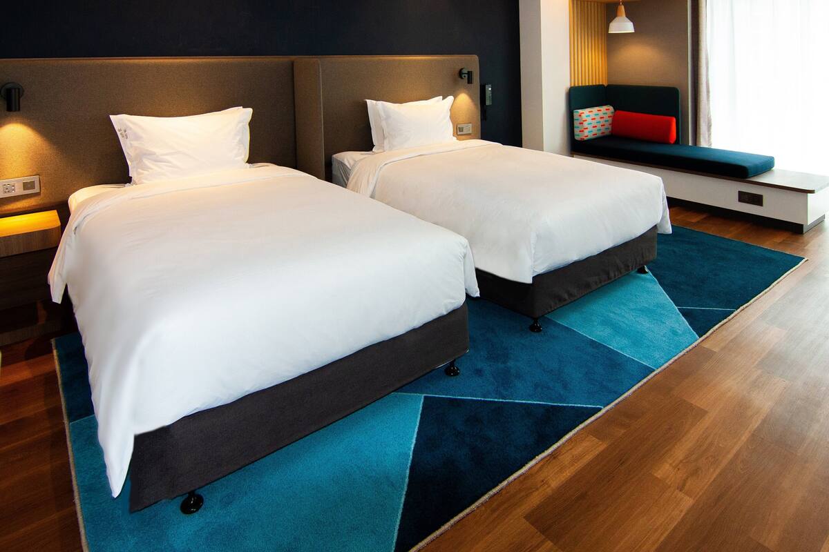 Habitación de hotel con dos camas, alfombra azul, suelo de madera, una banca con cojines de colores y una ventana con persianas.