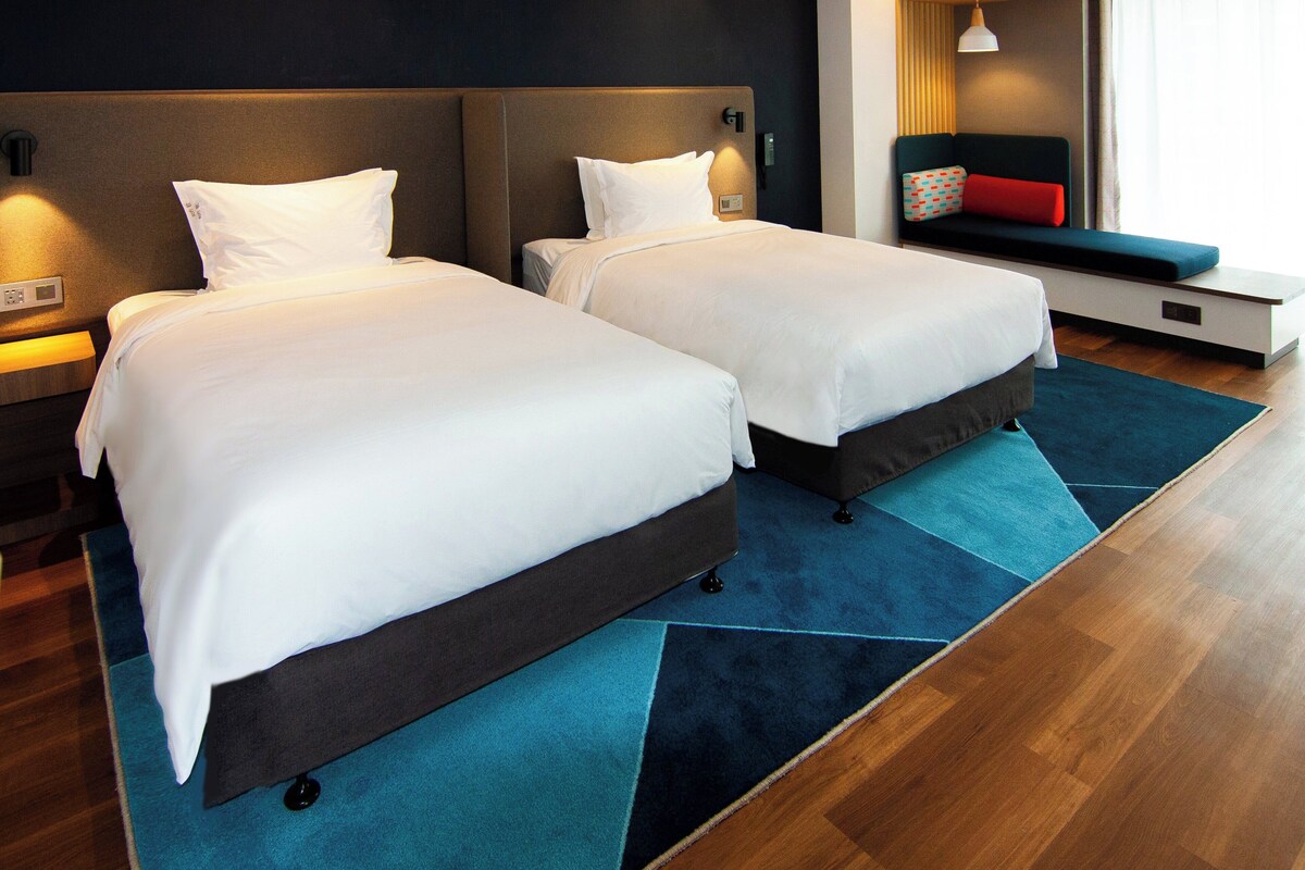 Habitación de hotel con dos camas, alfombra azul, suelo de madera, una banca con cojines de colores y una ventana con persianas.