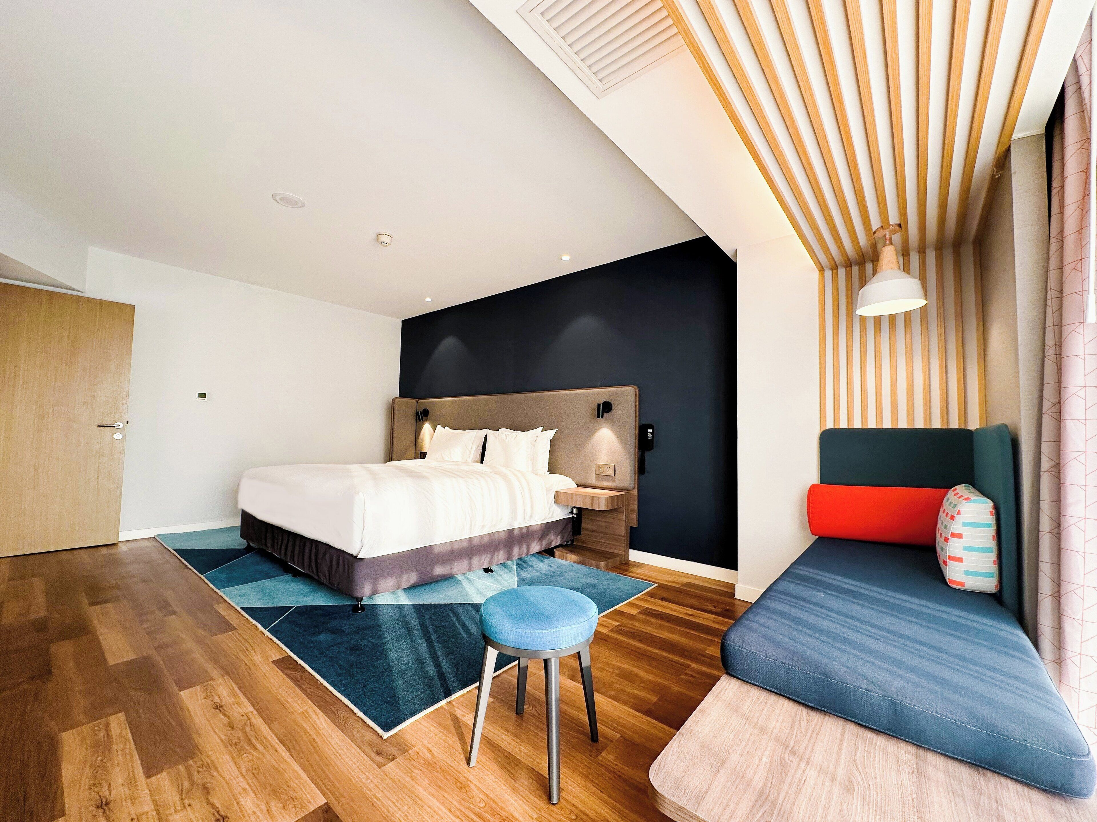Ein modernes Hotelzimmer mit einem großen Bett, einem Nachttisch, einem blauen Sessel und einer Couch mit bunten Kissen.