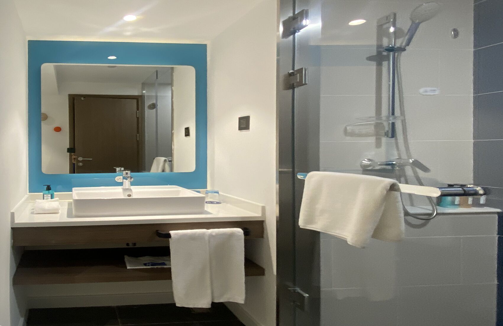 A modern bathroom with a white sink, a mirror with a blue frame, a glass shower enclosure, and a towel rack.