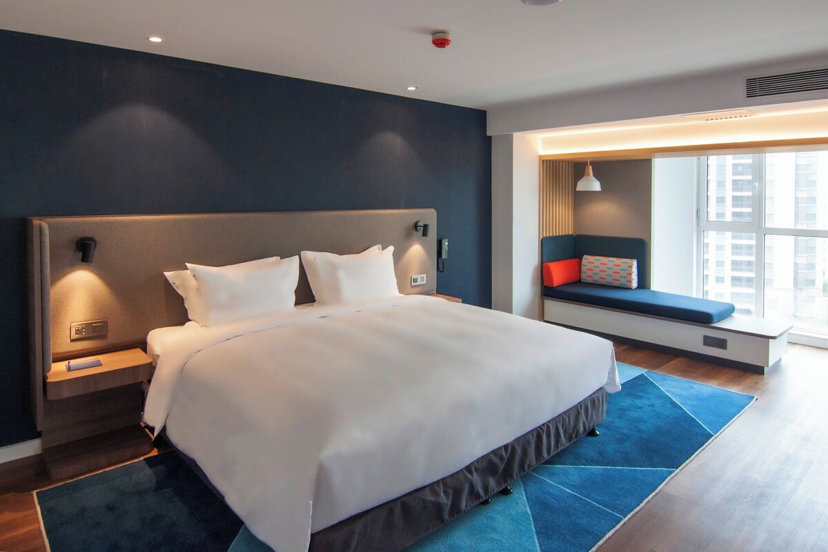Habitación de hotel moderna con una cama grande, mesitas de noche, un banco empotrado con cojines y un amplio ventanal con vistas a la ciudad.