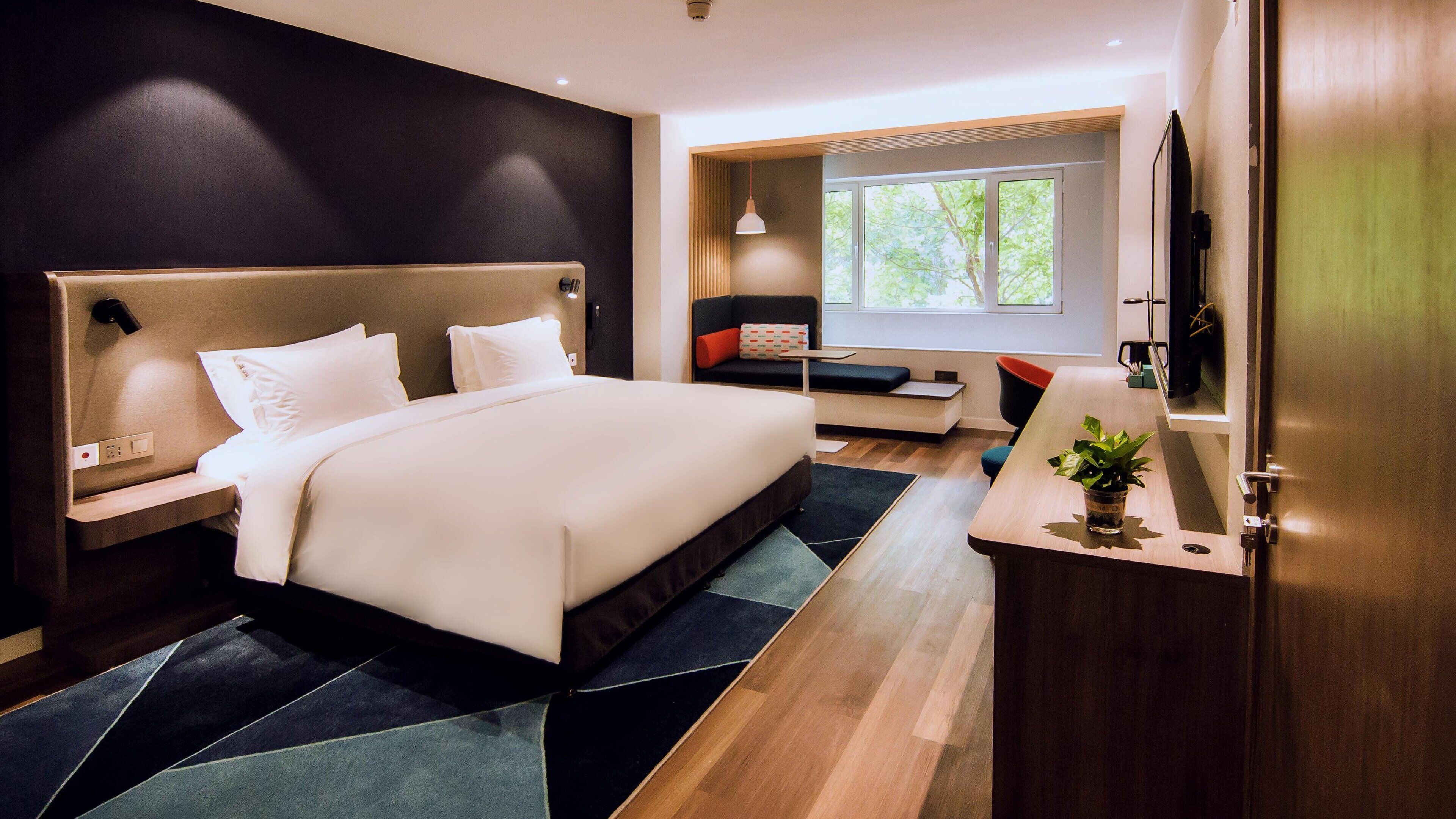 Una habitación de hotel moderna con una cama grande, un escritorio con una planta, una zona de estar y un ventanal amplio.