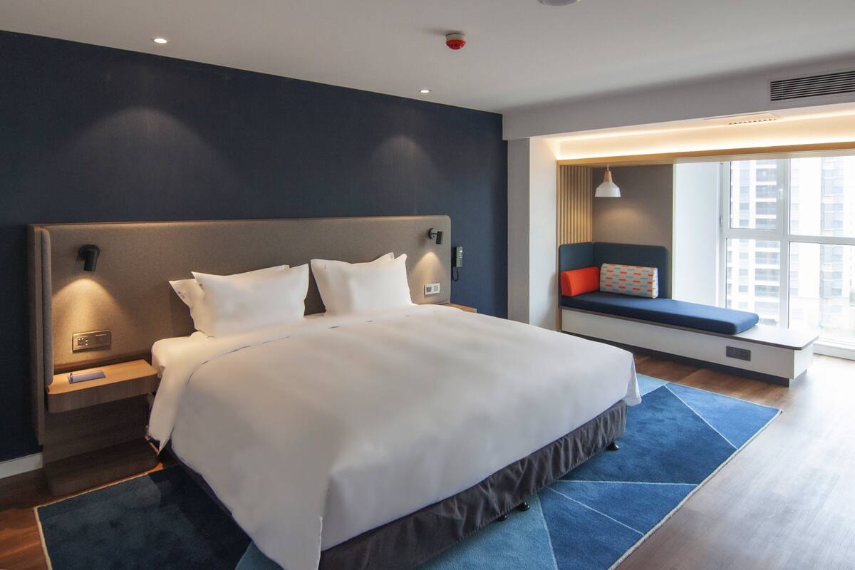 Habitación de hotel moderna con una cama grande, mesitas de noche, un banco empotrado con cojines y un amplio ventanal con vistas a la ciudad.