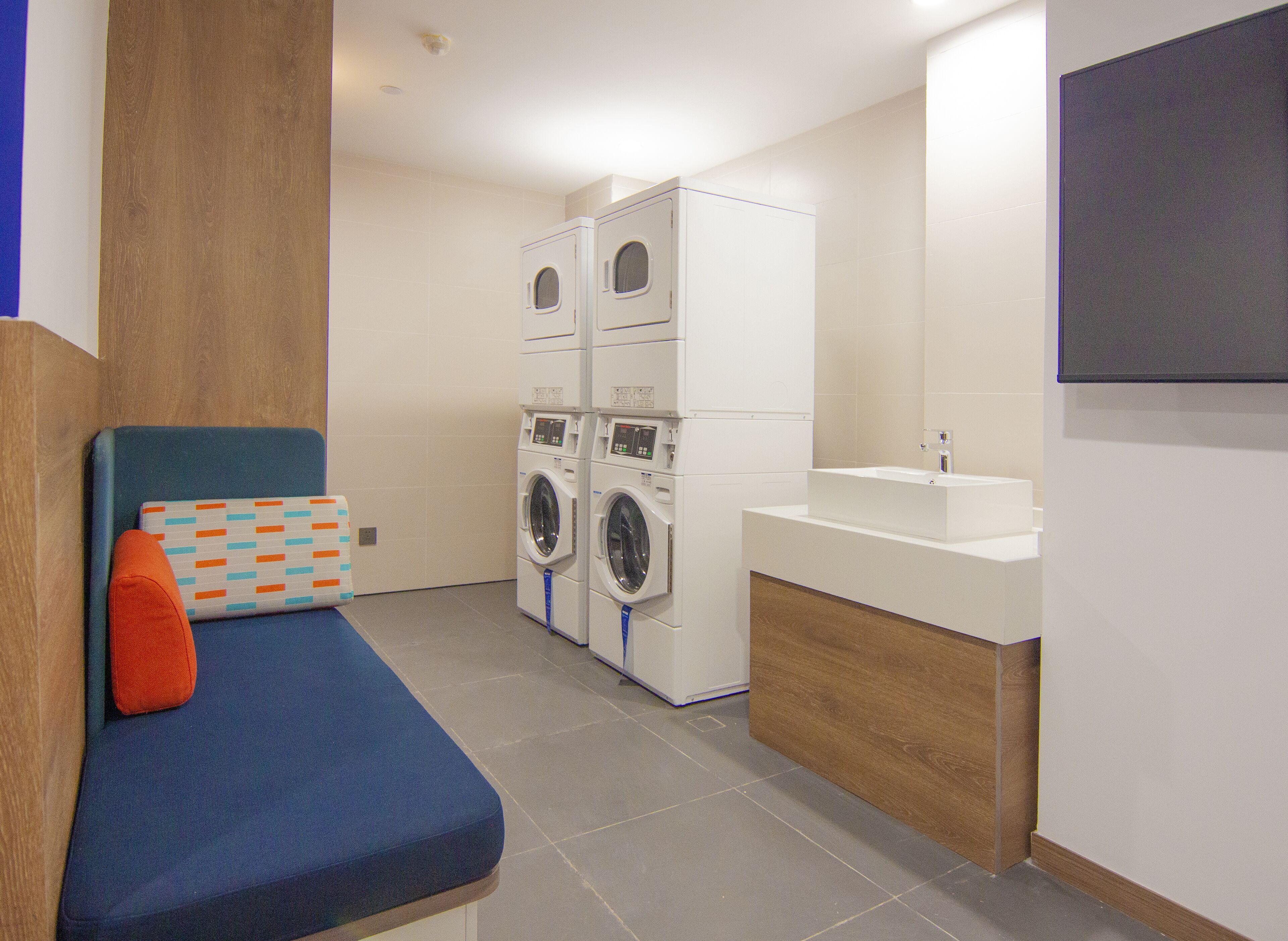 Une buanderie avec des lave-linge et des sèche-linge superposés, un banc bleu agrémenté d’un coussin à motifs et un évier blanc avec un meuble en bois.