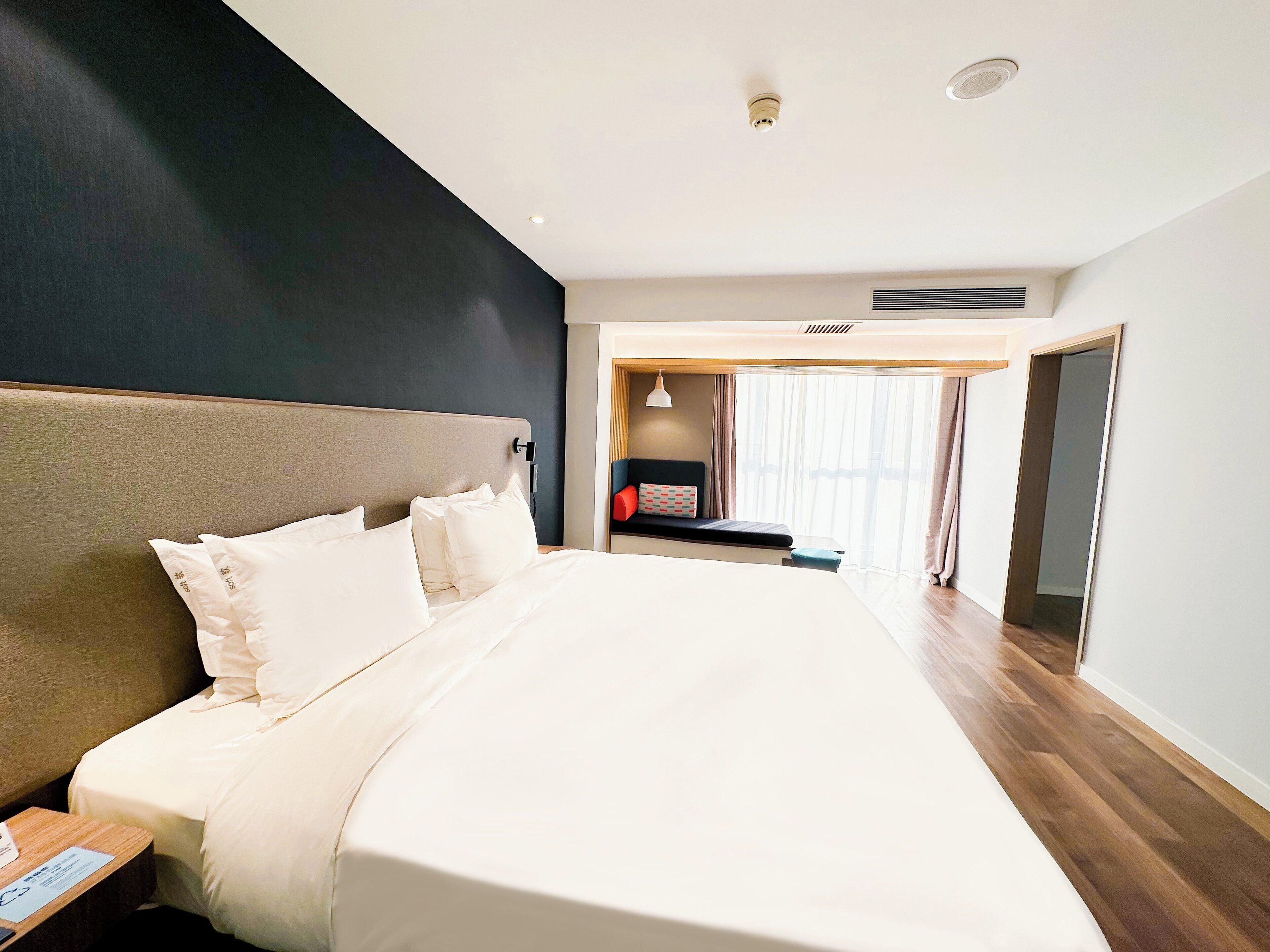 Una habitación de hotel moderna con una cama grande, una mesita de noche, cortinas en la ventana y una puerta que da a otra habitación.