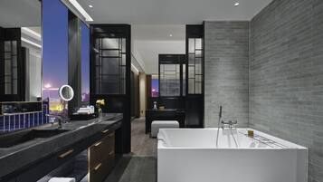 Un baño moderno con una bañera blanca grande, muebles de madera oscura y vista a la ciudad a través de un amplio ventanal.