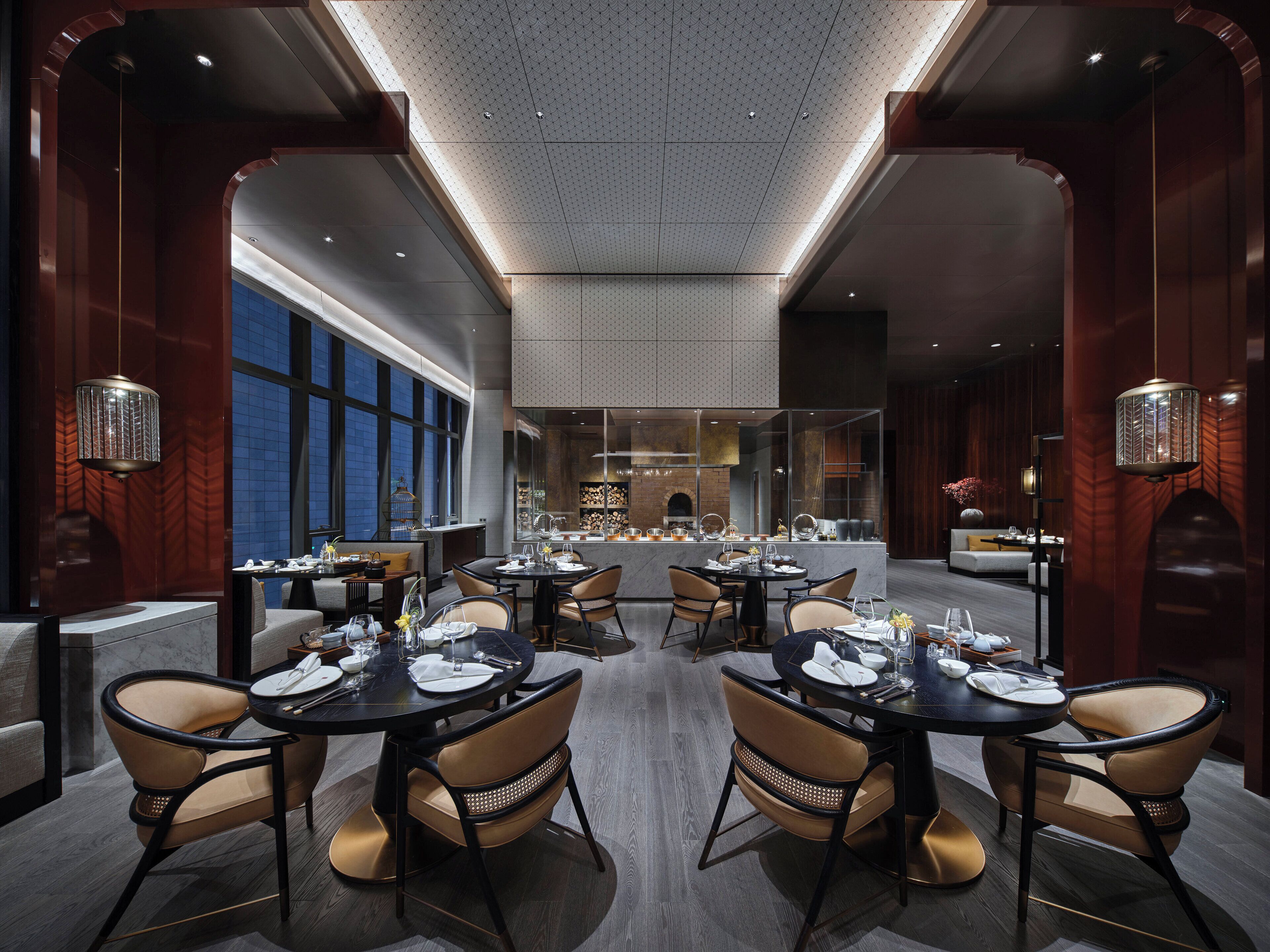 Uma sala de jantar moderna com várias mesas e cadeiras, teto alto com padrão geométrico e grandes janelas que permitem entrada de luz natural.