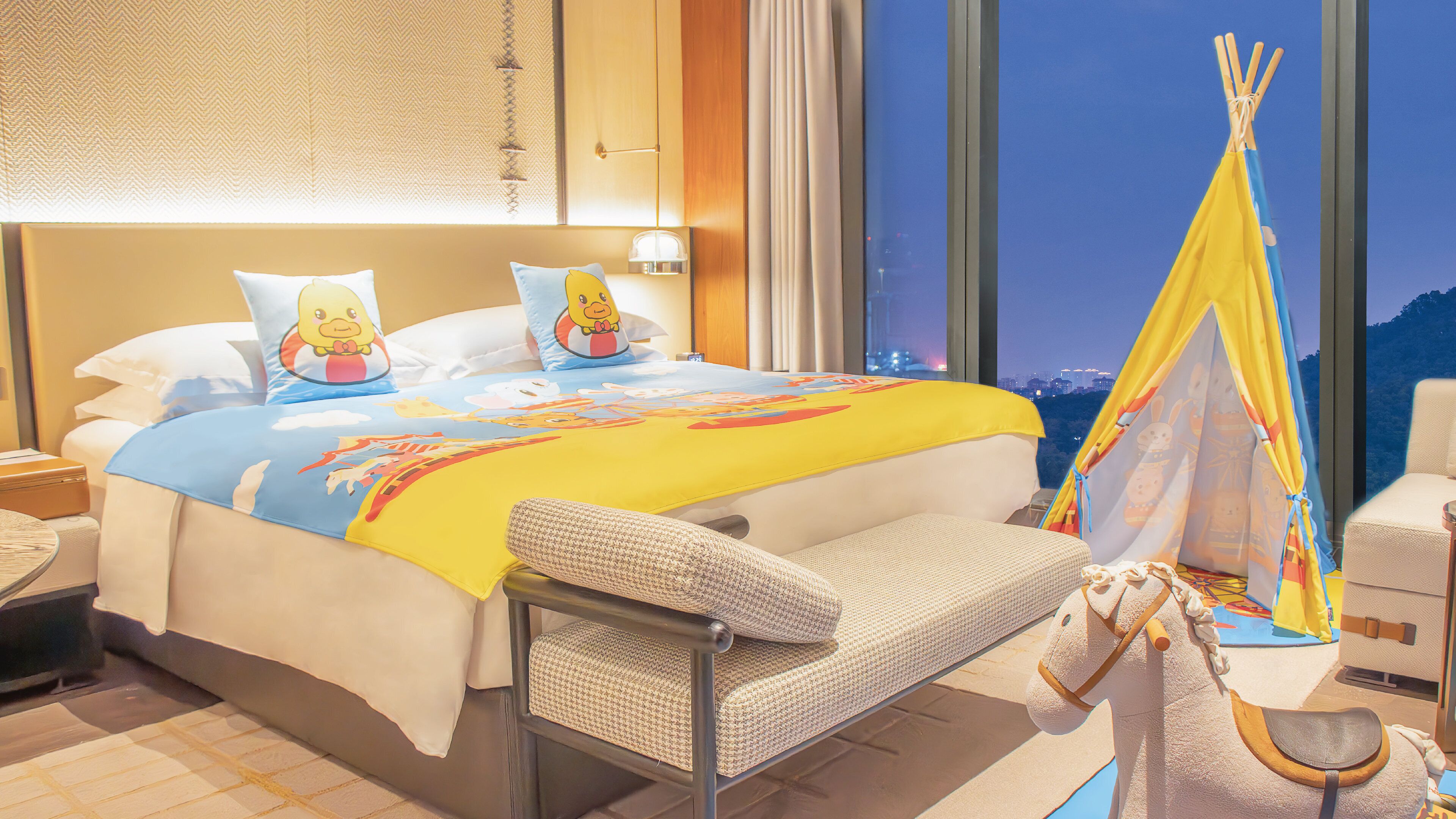 A hotel room with a large bed, a chair, a small table, and a view of the city at night.