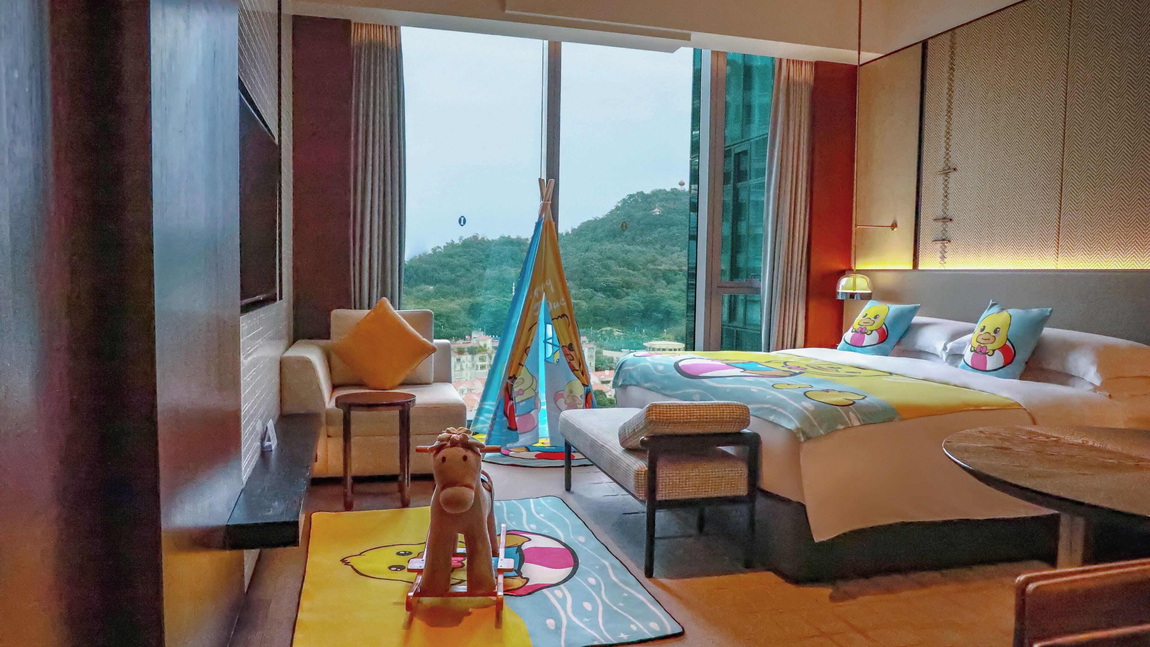 A hotel room with a bed, a sofa, a small table, and a view of greenery outside.