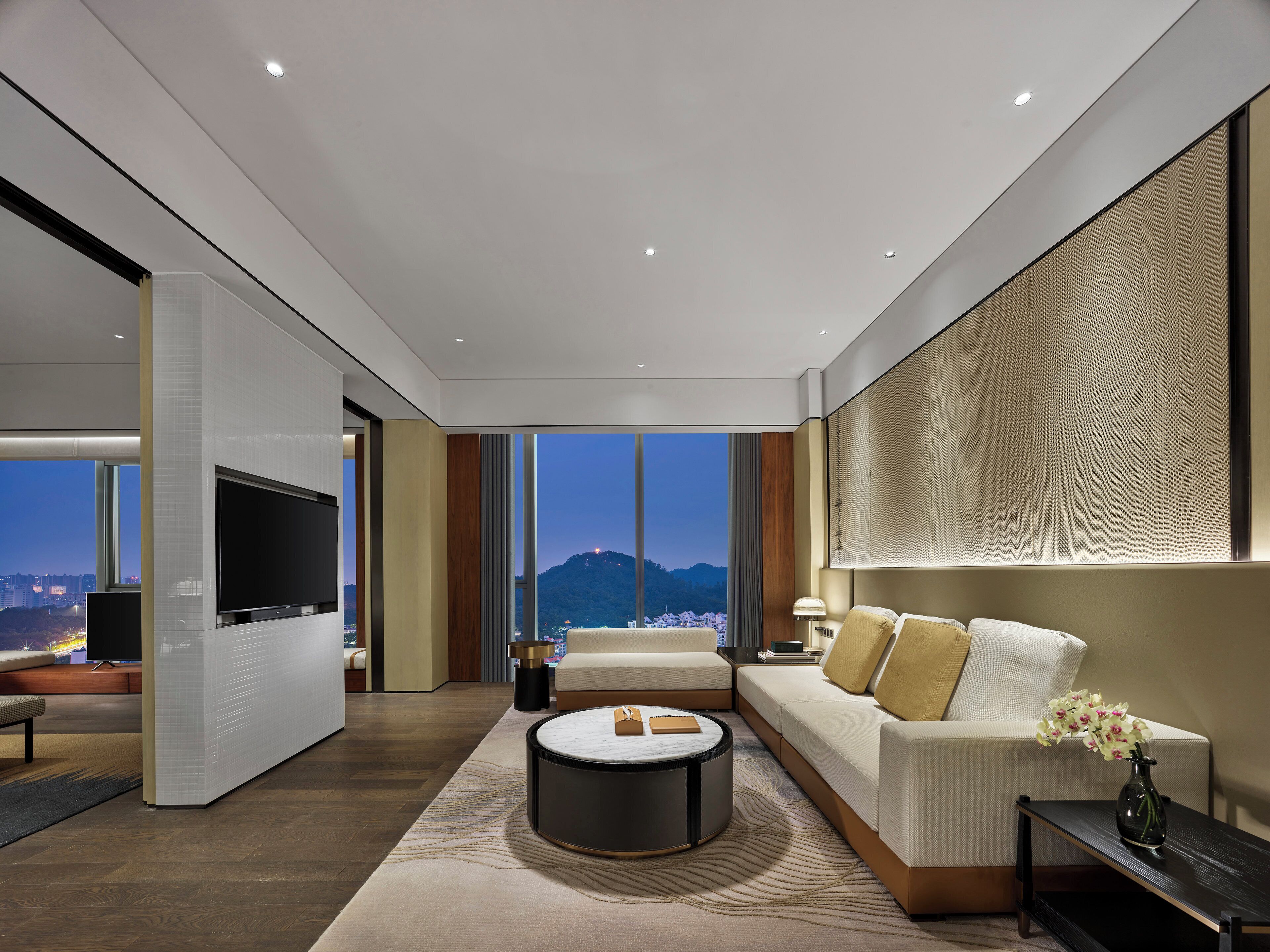 Una sala de estar moderna con un televisor grande, un sofá, una mesa de centro y vistas a un paisaje urbano al anochecer.