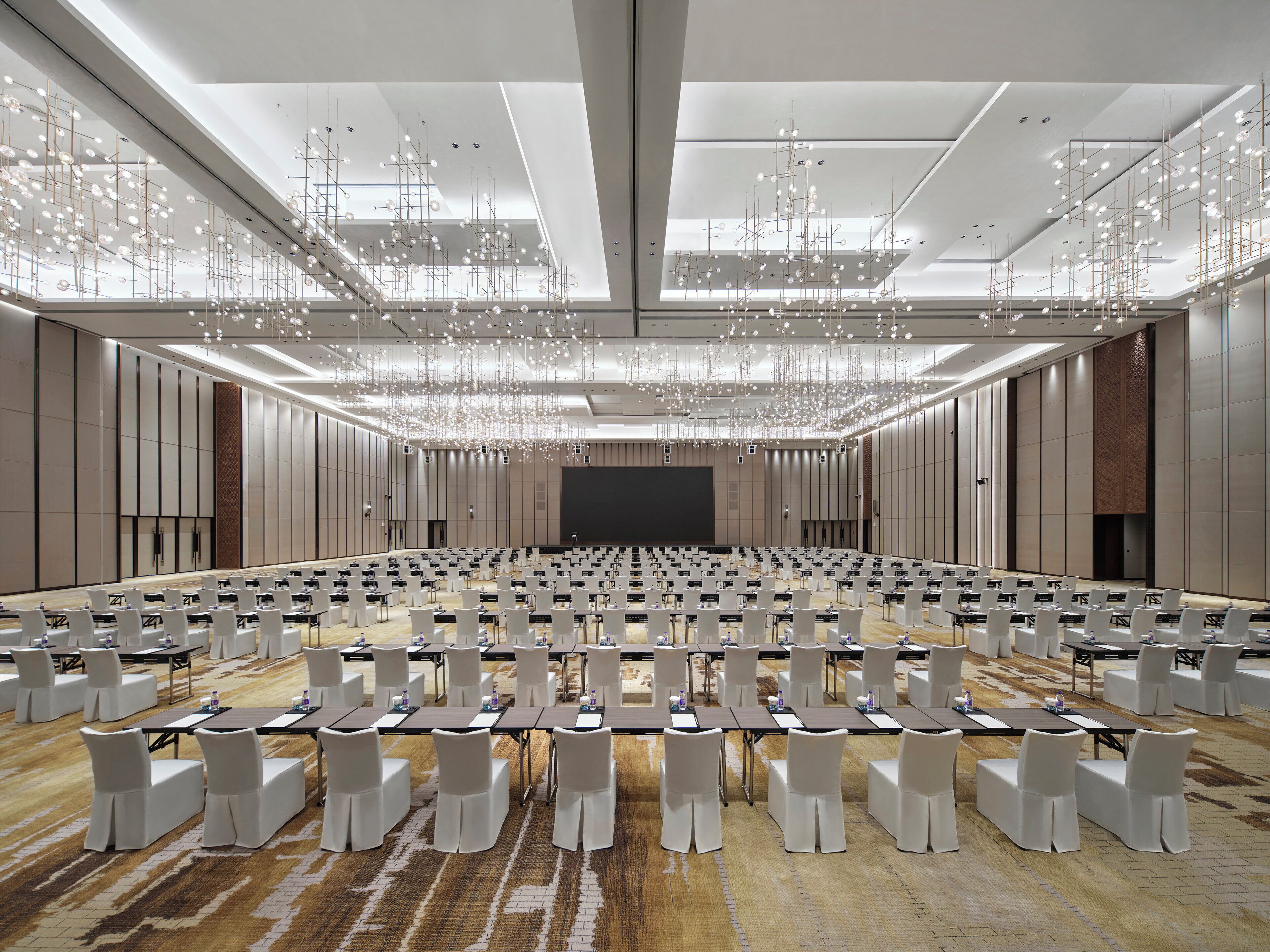 A large conference room with rows of tables and chairs, a stage, and a modern chandelier.