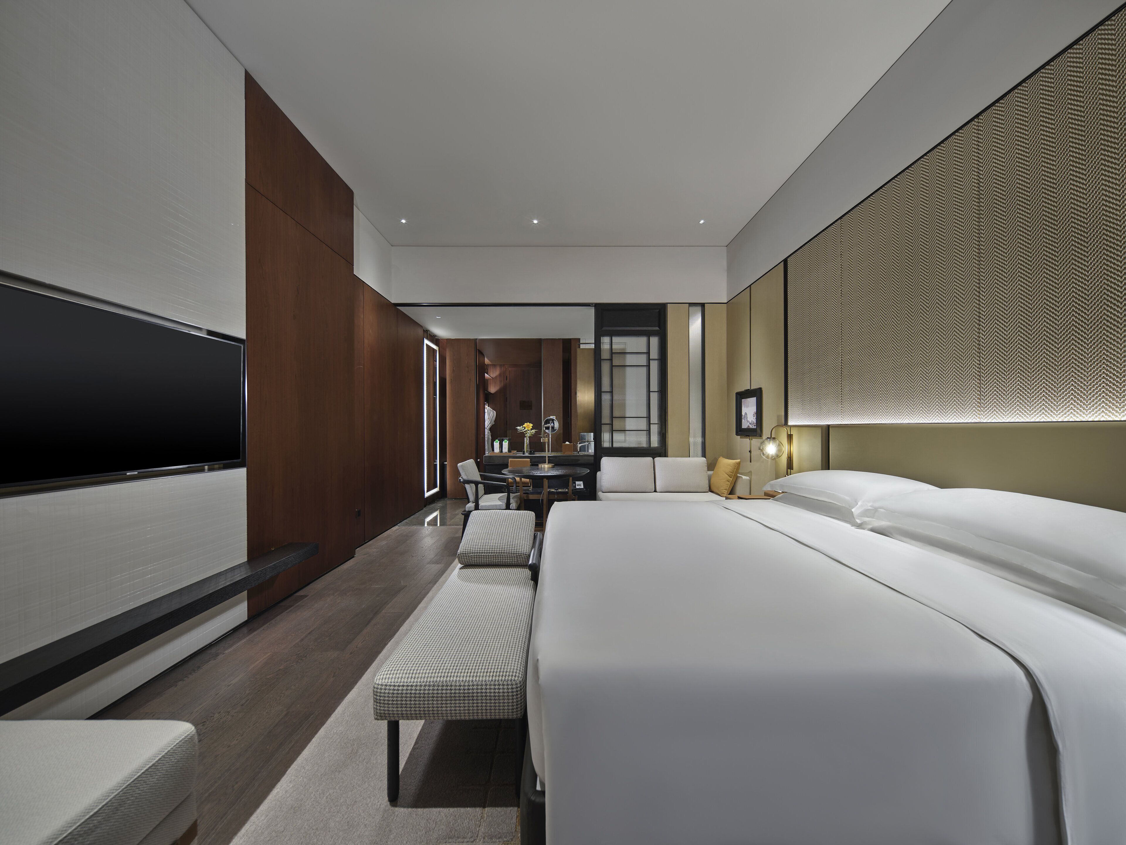 A modern hotel room with a large bed, a flat-screen TV, a dining area, and a view of the city.