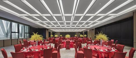 Un amplio salón de conferencias con mesas dispuestas para un evento formal, destacando un diseño moderno del techo y ventanas grandes.