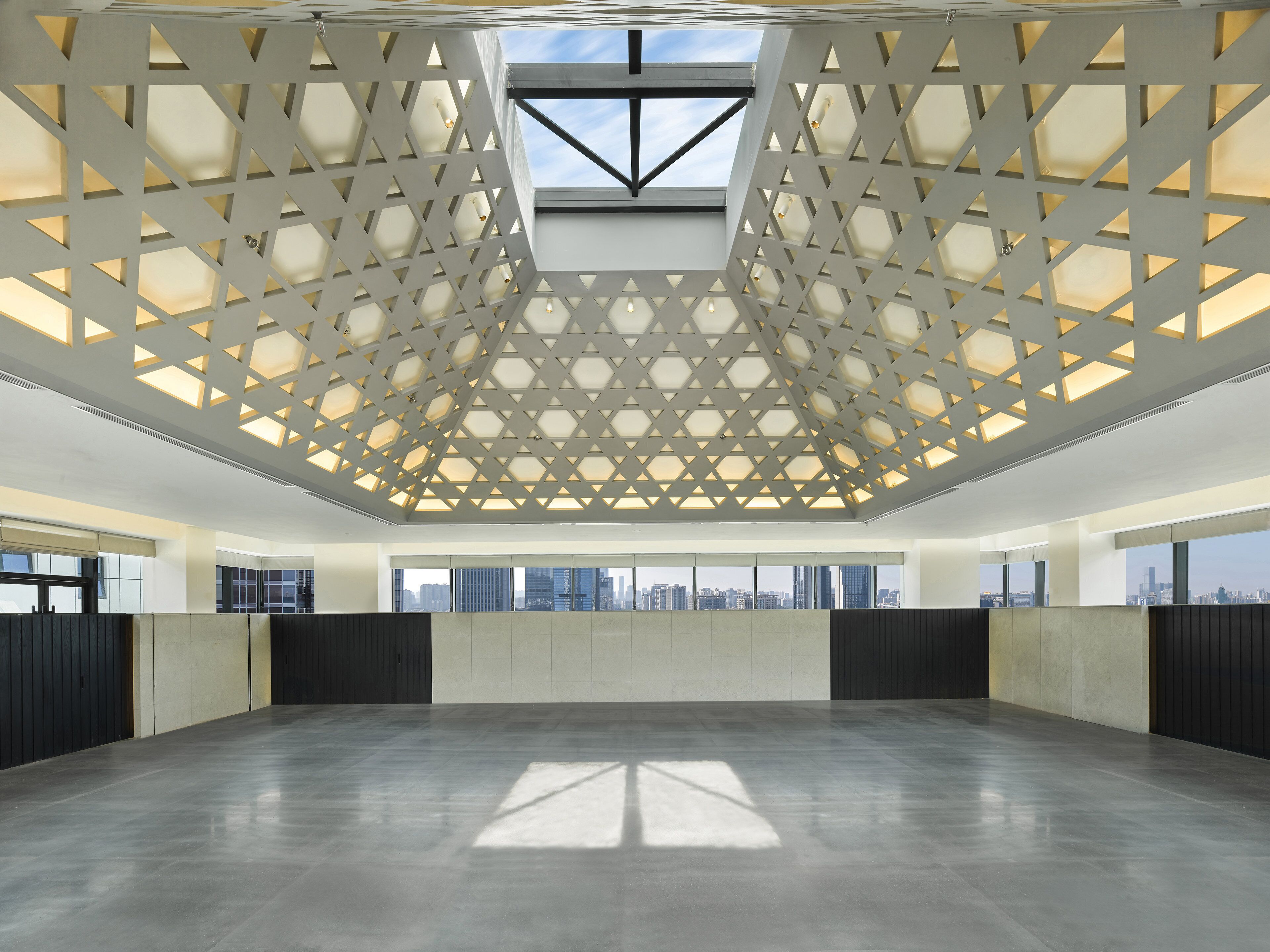 A spacious interior with a high ceiling, geometric patterned ceiling, large windows, and a view of the city skyline.
