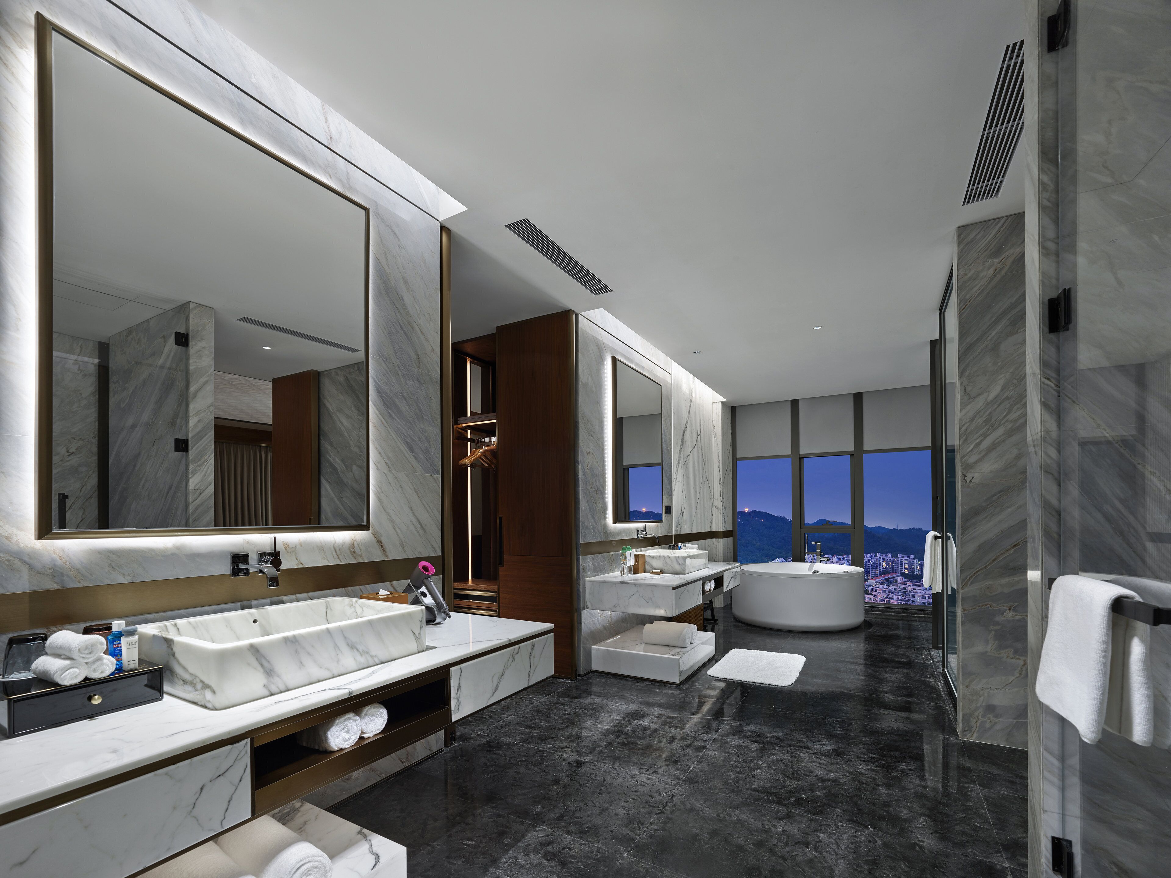 Un baño moderno con un espejo grande, lavamanos de mármol, una bañera y vistas a un paisaje urbano.